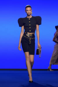 Model walks a blue-lit runway in a black two-piece outfit: cropped top with oversized puff sleeves, gold buttons, matching shorts, and a gold belt with chain accents, holding a small black purse with a gold chain.