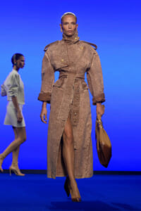 Model walks the runway in a textured tan belted trench coat with oversized shoulders and gold buttons, carrying a tan handbag against a blue background.