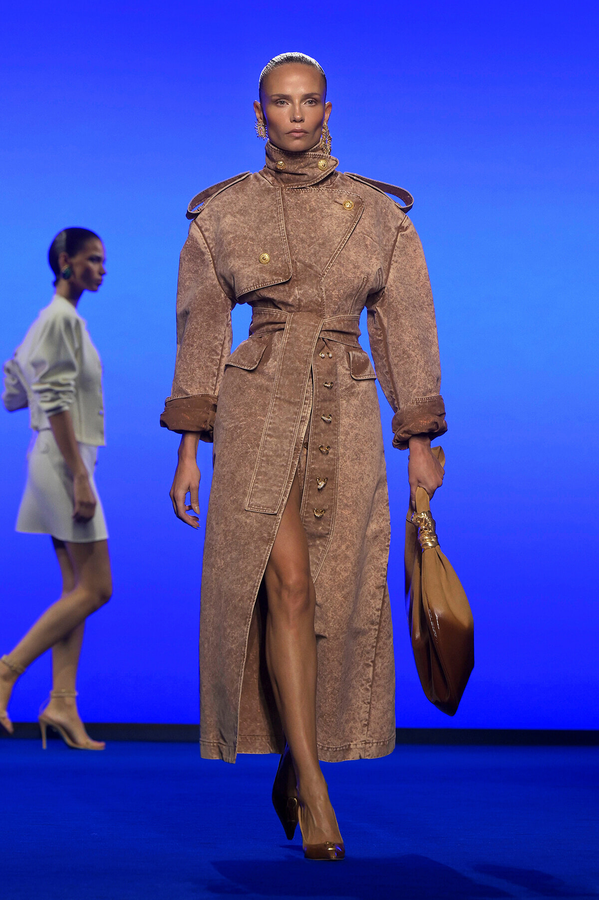 Model walks the runway in a textured tan belted trench coat with oversized shoulders and gold buttons, carrying a tan handbag against a blue background.