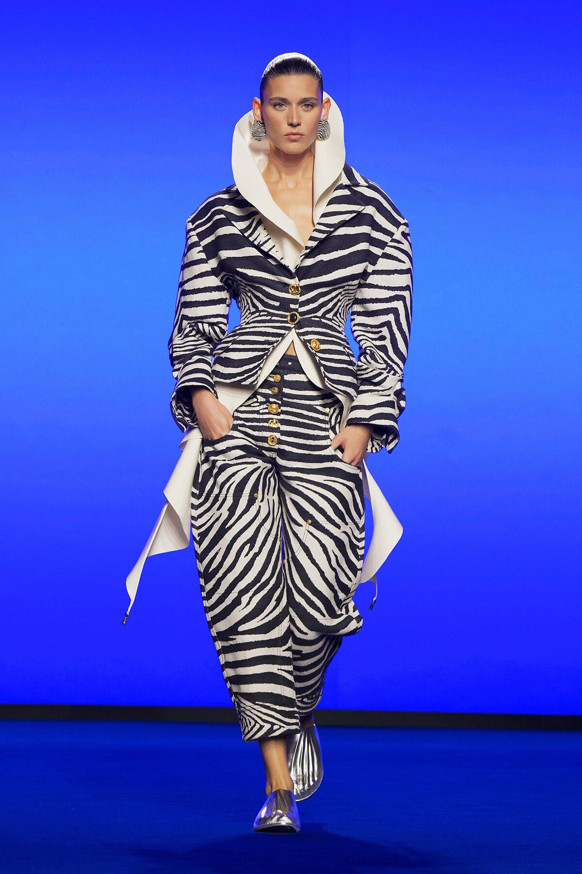 Model on a runway wearing a bold zebra-print suit with an oversized white collar, gold buttons, and metallic shoes against a blue backdrop.
