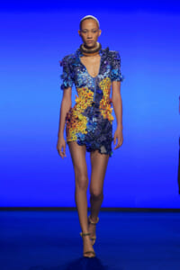 Model walking a runway in a short, multicolored floral dress with blue and yellow petals, paired with gold accessories and heels against a blue backdrop.