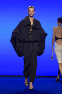 Model walks the runway in a voluminous black coat with oversized sleeves, gold zipper and belt, against a blue backdrop.