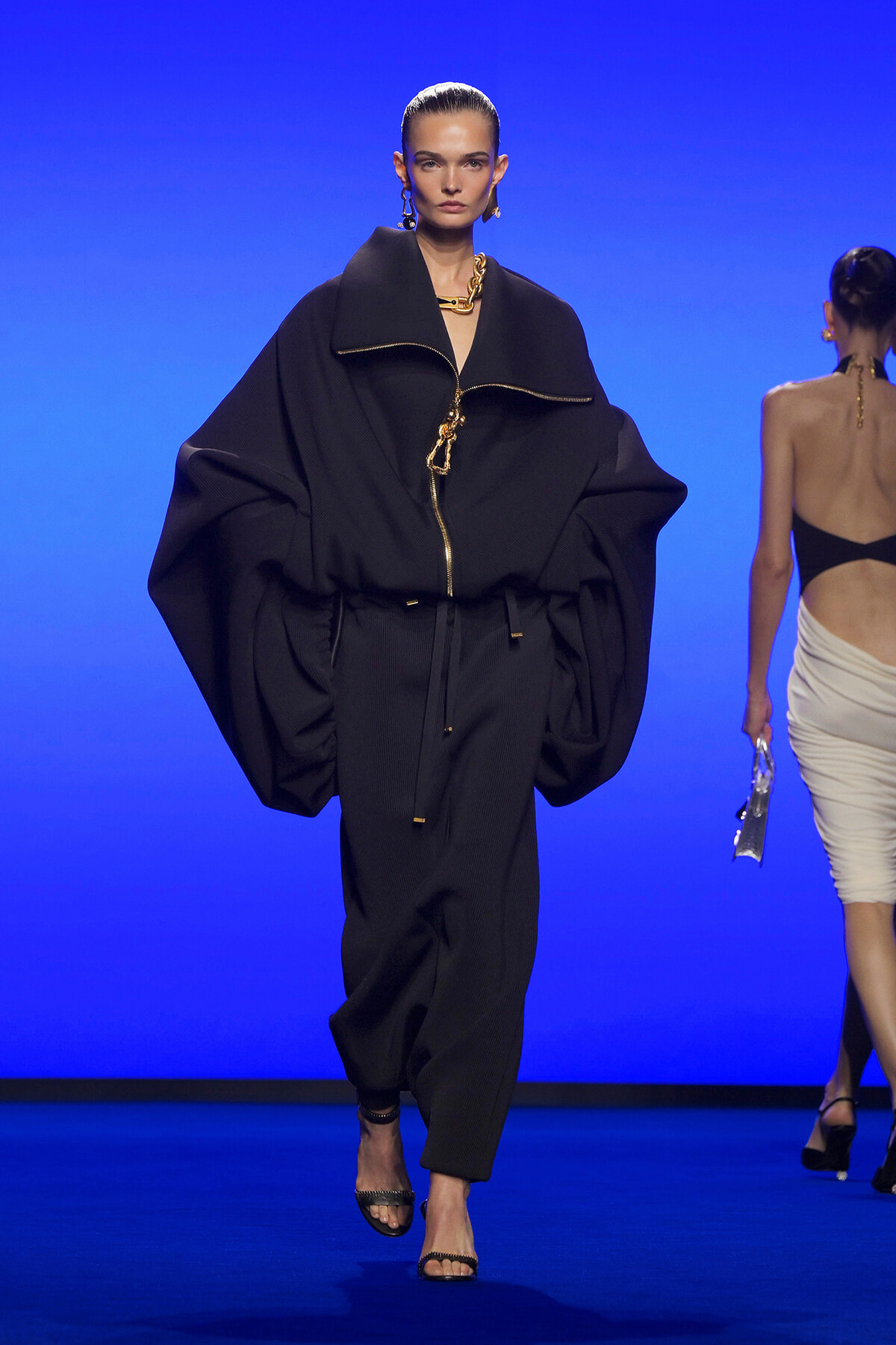 Model walks the runway in a voluminous black coat with oversized sleeves, gold zipper and belt, against a blue backdrop.