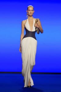 Model in a white draped gown with a black waist corset, holding a zebra-striped clutch against a blue runway backdrop.