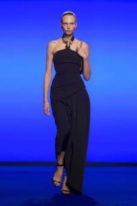 Model walking a runway in a black halter jumpsuit with a gold statement necklace against a blue backdrop.