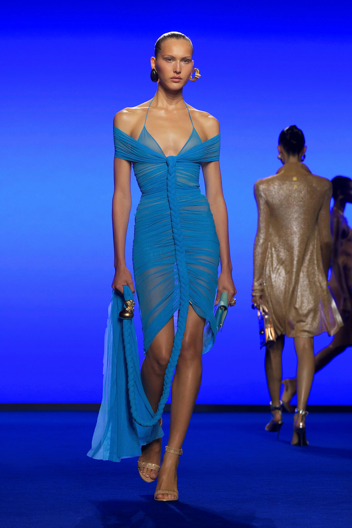 Model walks the runway in a turquoise off-shoulder, ruched midi dress with a front knot, holding a matching scarf and clutch against a blue backdrop.
