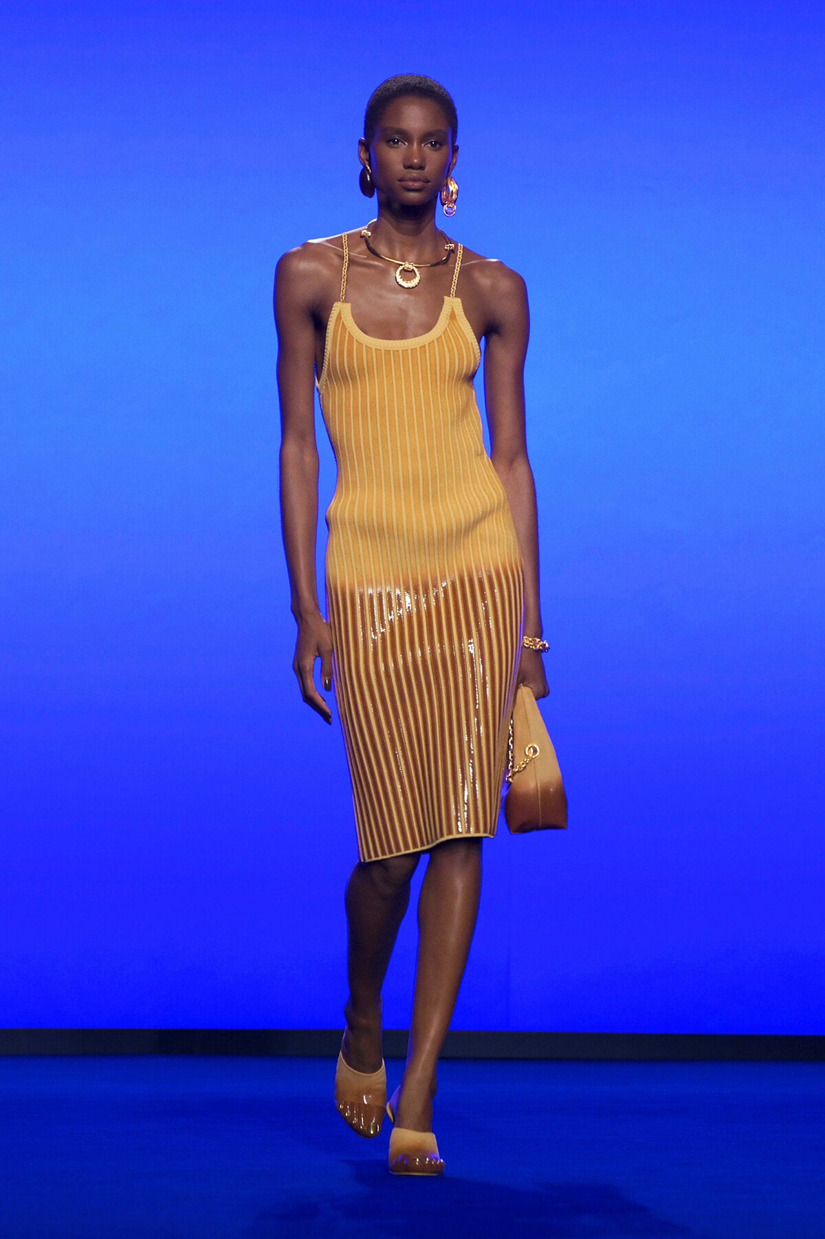 Model walking a fashion runway in a mustard-yellow striped dress with a sheer lower half, gold jewelry, and a tan handbag against a blue backdrop