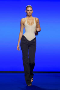 Model walking the runway in a beige corset-style top and dark jeans, carrying a brown bag with a chain strap against a blue backdrop.
