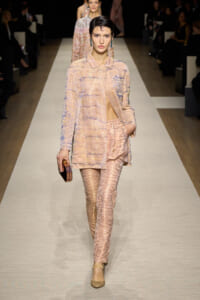 Model walks the runway in a peach sequined suit with a matching coat, patterned pants, and a geometric clutch in hand.