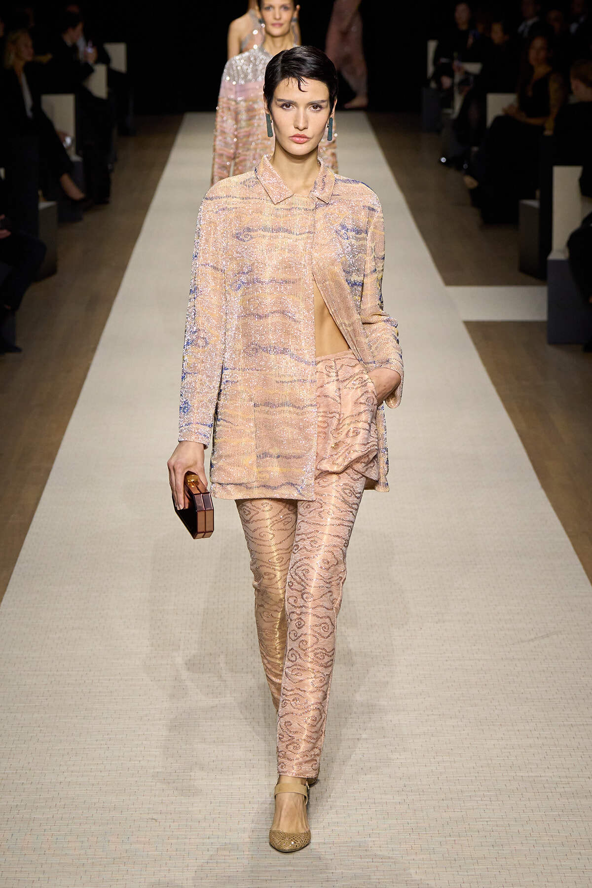 Model walks the runway in a peach sequined suit with a matching coat, patterned pants, and a geometric clutch in hand.