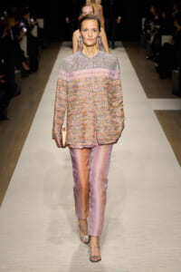 Model struts down a fashion-show runway in a pale pink patterned jacket, matching satin pants, and glittery sandals, clutch in hand.