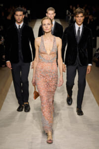 Female model in a peach beaded halter gown walking the runway, flanked by three male models in black velvet blazers.