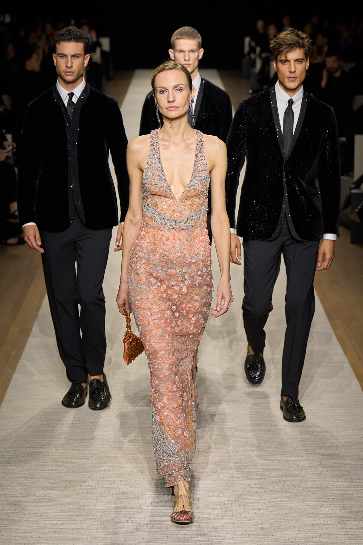 Female model in a peach beaded halter gown walking the runway, flanked by three male models in black velvet blazers.