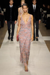 Model walks the runway in a pink sequined deep-V gown with a pink clutch, surrounded by other models in black outfits.