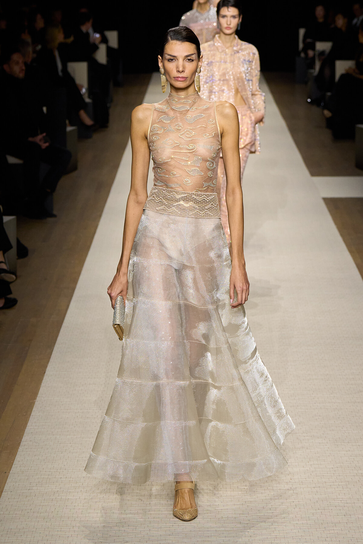 Model on a runway wearing a sheer beige lace sleeveless bodice and a flowing, light‑metallic skirt, accessorized with a small clutch and gold earrings.