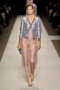 Model walking a fashion runway wearing a light blue embellished cardigan over a sheer pink lace jumpsuit, holding a small clutch.