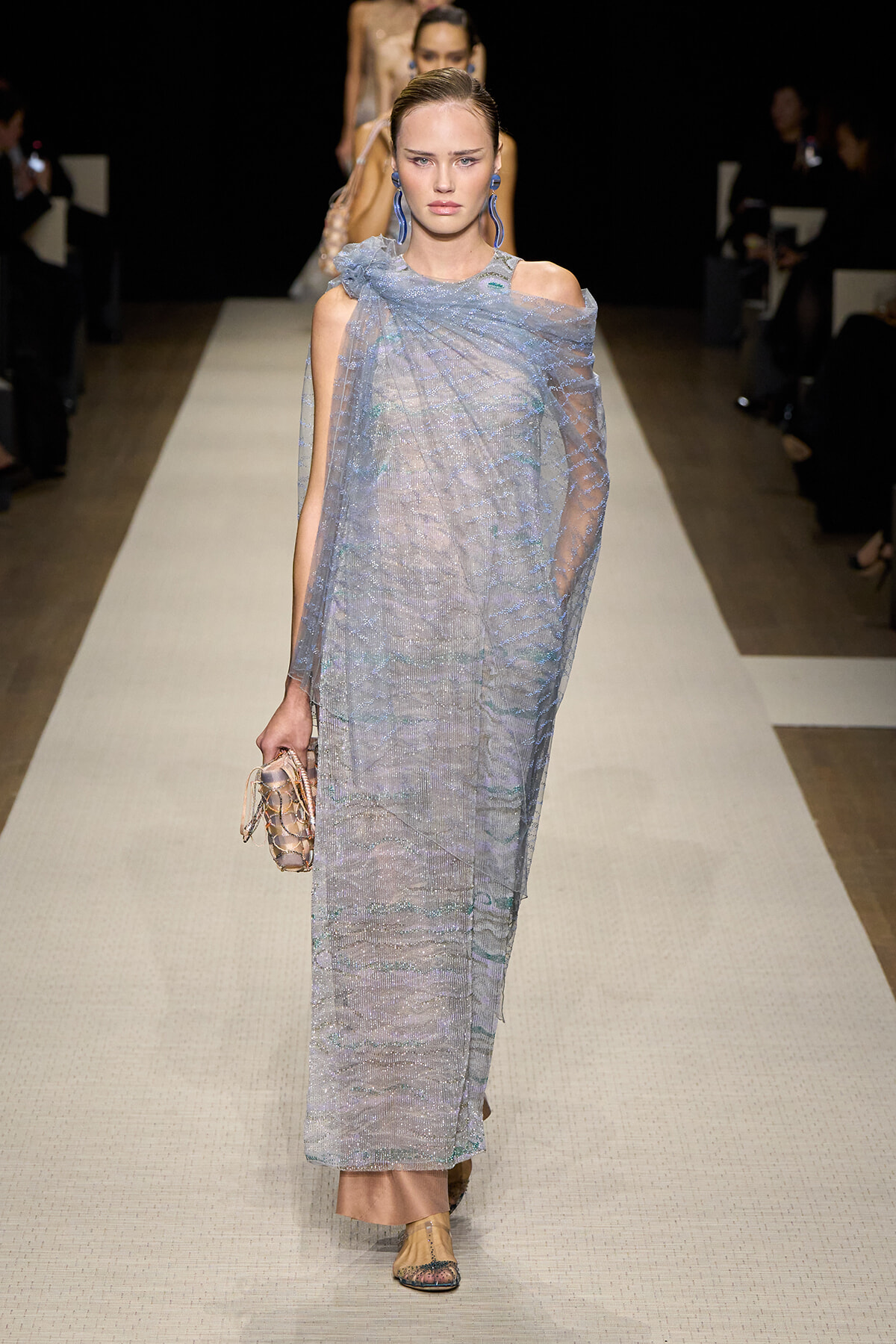 Model on a fashion show runway wearing a light blue, sheer, shimmering full-length dress with an off-shoulder overlay, holding a small woven clutch.