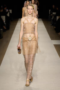 Model walking the runway in a champagne sequin crop top and matching sheer sequined pants with a draped belt and gold accessories.