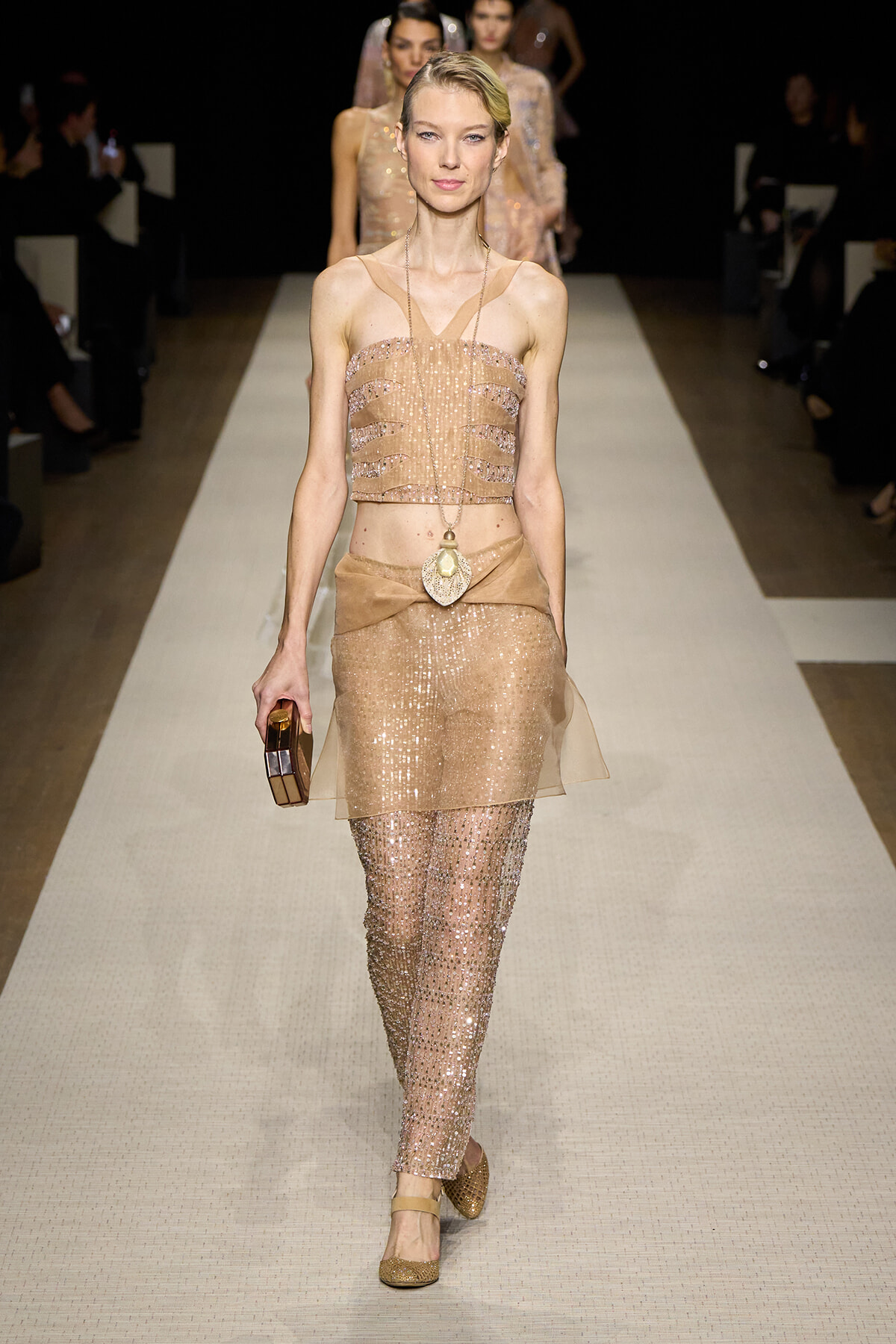 Model walking the runway in a champagne sequin crop top and matching sheer sequined pants with a draped belt and gold accessories.