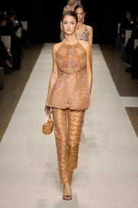 Model walking a runway in peach-toned, beaded cape top and patterned matching pants with a small handbag and matching jewelry.