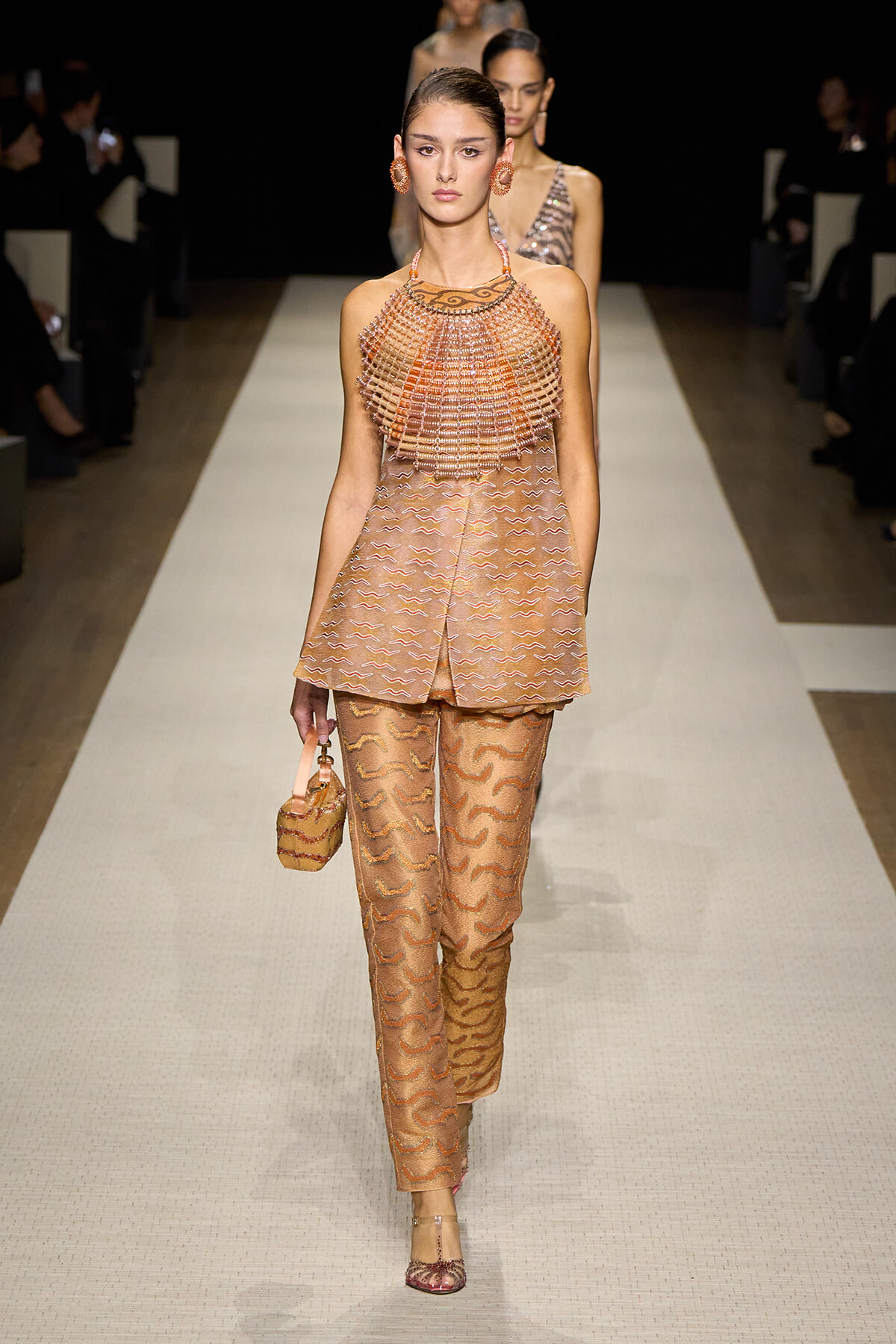 Model walking a runway in peach-toned, beaded cape top and patterned matching pants with a small handbag and matching jewelry.