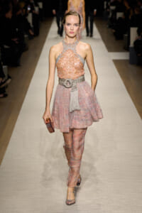 Model walks the runway in a blush pink sequined halter dress with a wide metallic belt, paired with pink patterned pants and a small burgundy clutch.