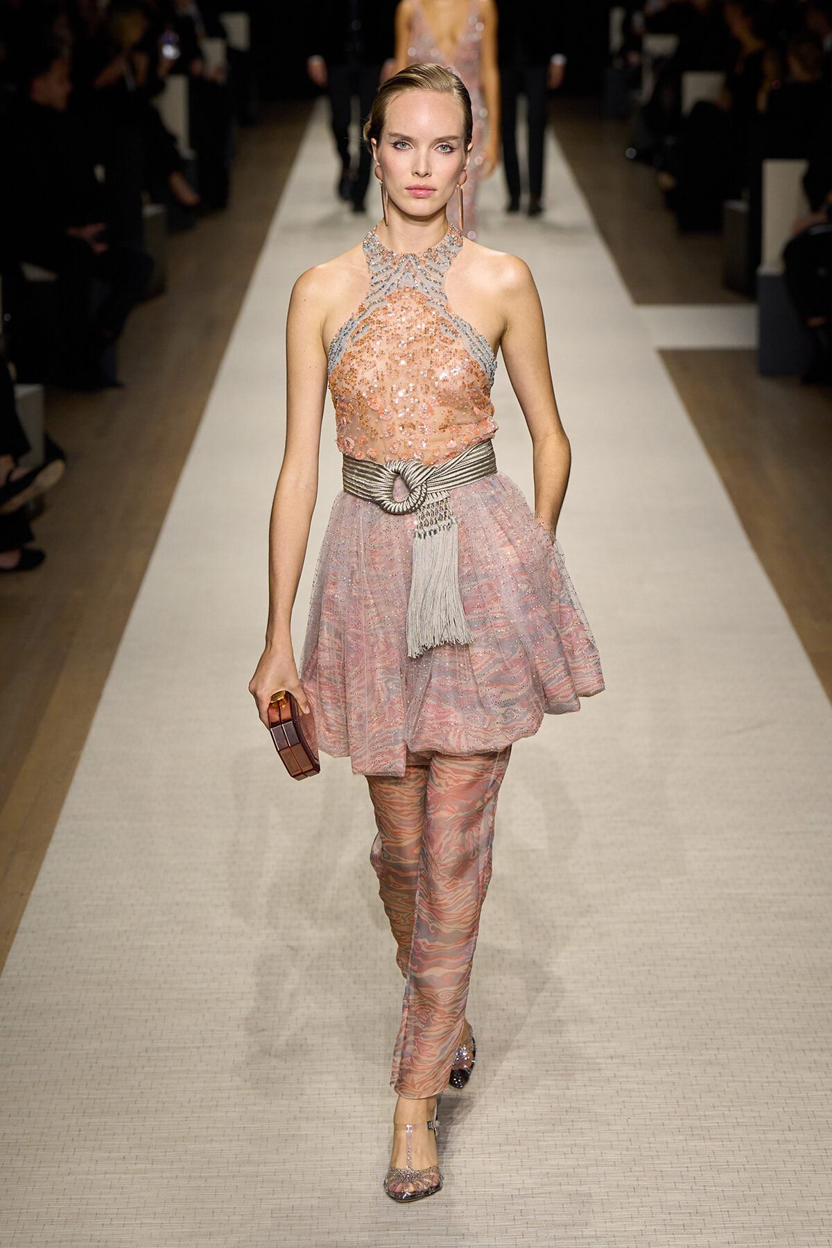 Model walks the runway in a blush pink sequined halter dress with a wide metallic belt, paired with pink patterned pants and a small burgundy clutch.