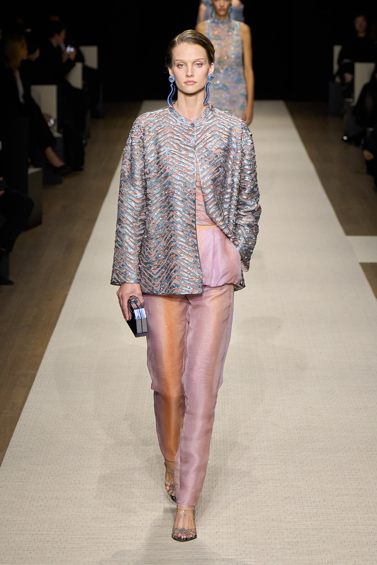 Model on a runway wearing a metallic chevron-patterned jacket with pink satin pants, blue earrings, and a small metallic box clutch.