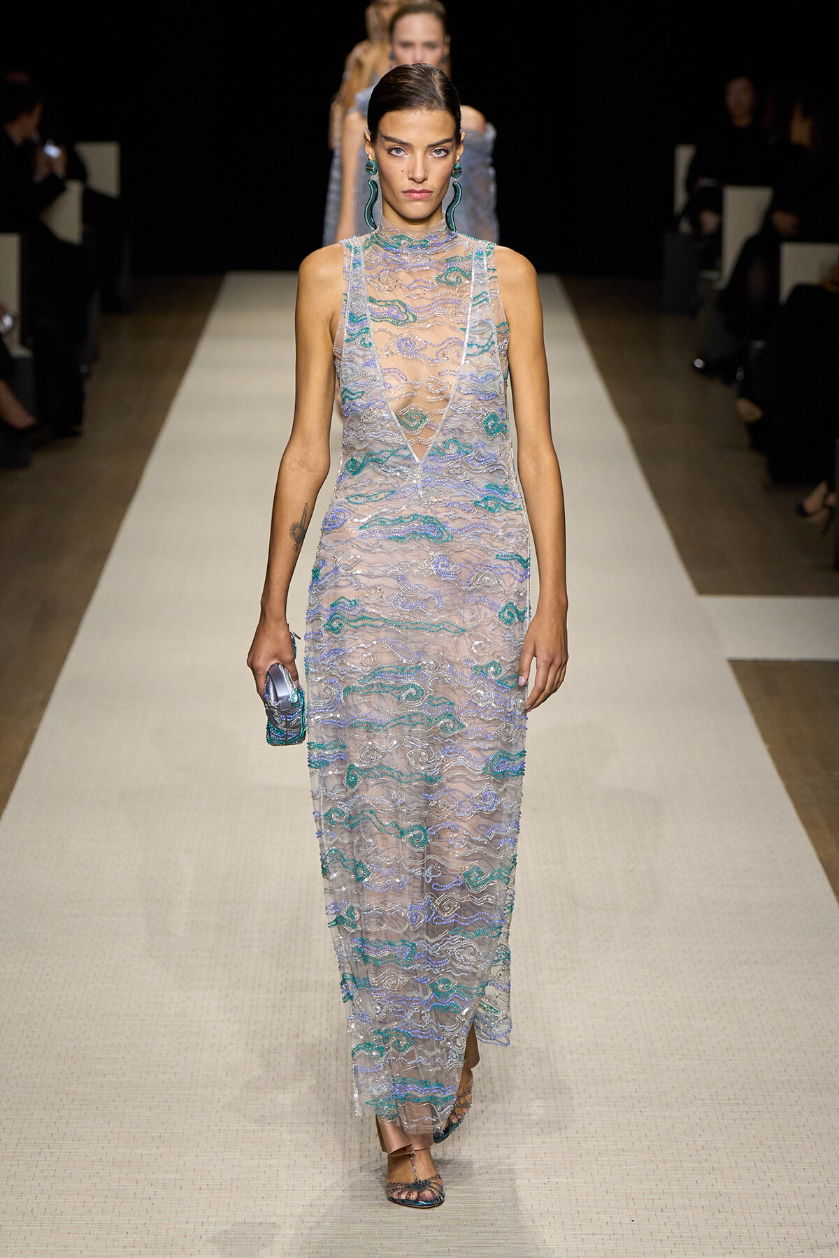 Model on a runway wearing a sheer, floor-length lace gown with teal and silver embroidery, carrying a small metallic clutch
