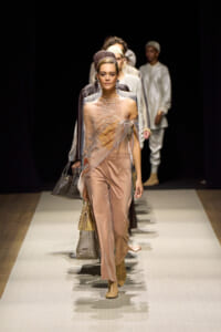 Model walks the runway in a peach satin jumpsuit with a sheer embroidered wrap, tan boots, and a headpiece; backstage-style crowd in the dark background.