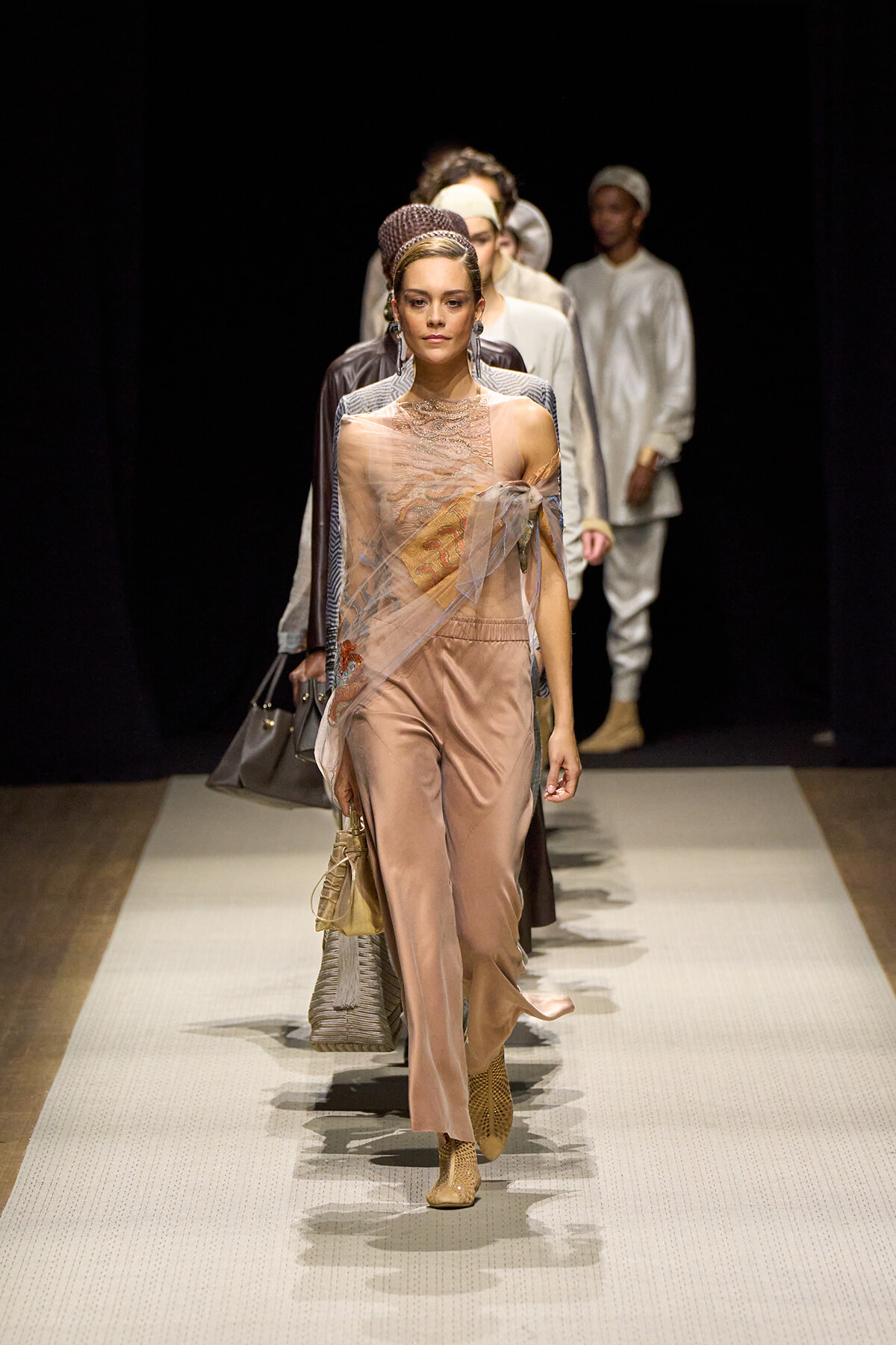 Model walks the runway in a peach satin jumpsuit with a sheer embroidered wrap, tan boots, and a headpiece; backstage-style crowd in the dark background.