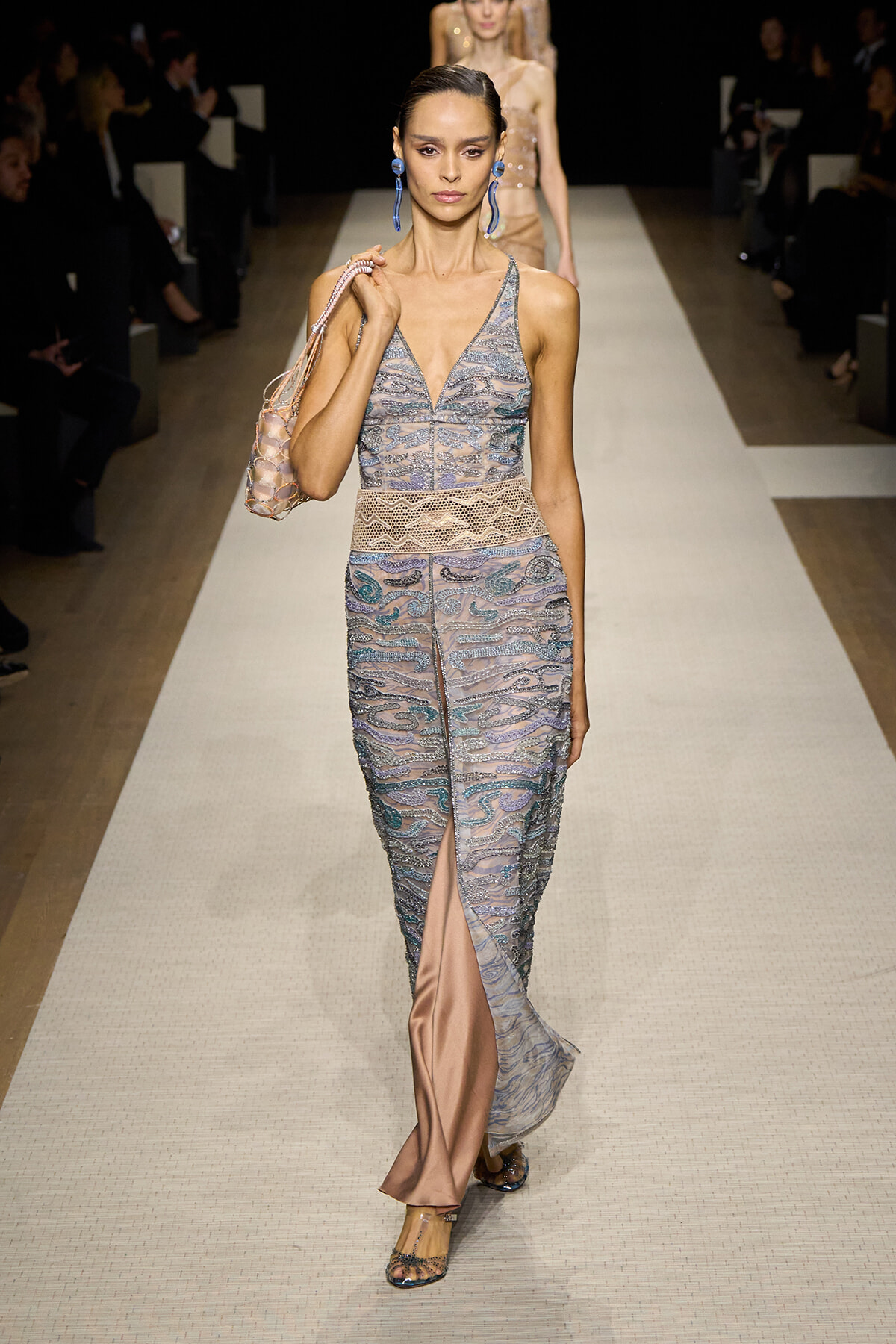 Model walks a runway wearing a sheer pastel lace gown with a V-neck, wide peach satin stripe at the bottom, and a front slit, carrying a netted bag and blue earrings.