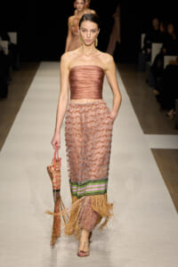 Female model walks a runway in a strapless pink-brown top and pink zigzag-patterned skirt with green-purple bands and fringed hem; she carries a woven bag.