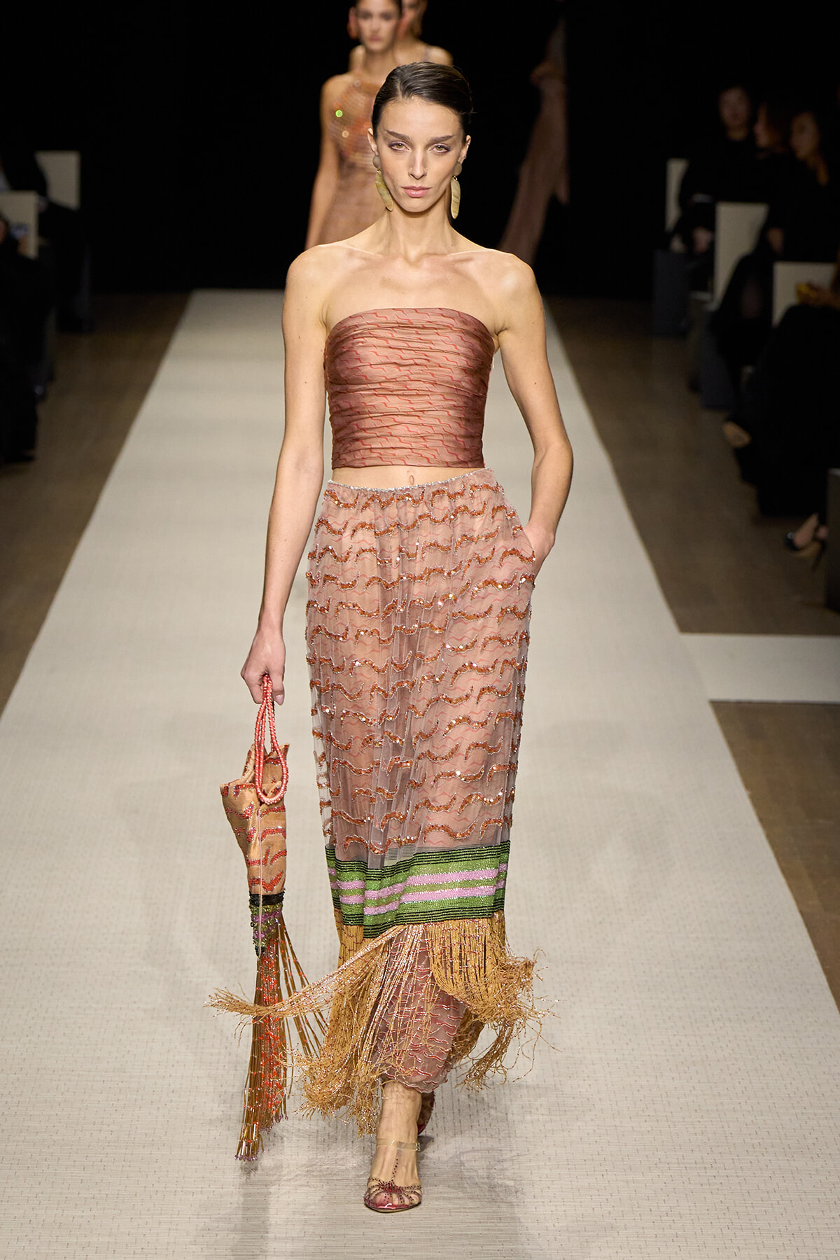 Female model walks a runway in a strapless pink-brown top and pink zigzag-patterned skirt with green-purple bands and fringed hem; she carries a woven bag.