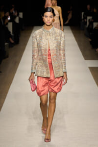 Model on a runway wearing a metallic beige jacquard jacket over pink satin bubble shorts, with a pink clutch and pink heels.