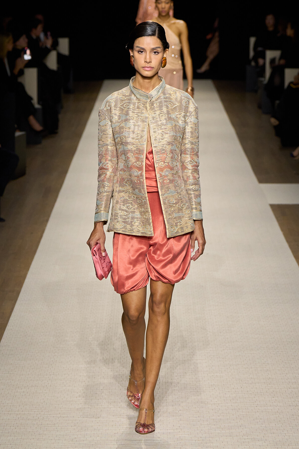 Model on a runway wearing a metallic beige jacquard jacket over pink satin bubble shorts, with a pink clutch and pink heels.