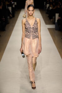 Model walking a runway in a peach satin jumpsuit with a patterned sleeveless vest and matching pants, carrying a small box clutch.