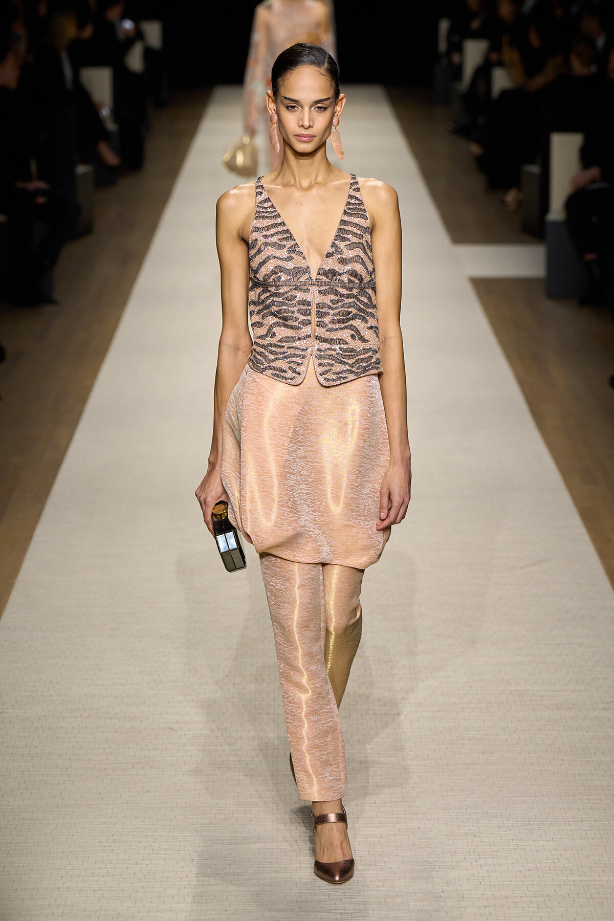 Model walking a runway in a peach satin jumpsuit with a patterned sleeveless vest and matching pants, carrying a small box clutch.