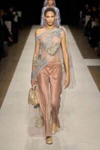 Model walks the runway in a sheer, embroidered cape over pale pink satin pants, paired with a metallic handbag and woven boots.