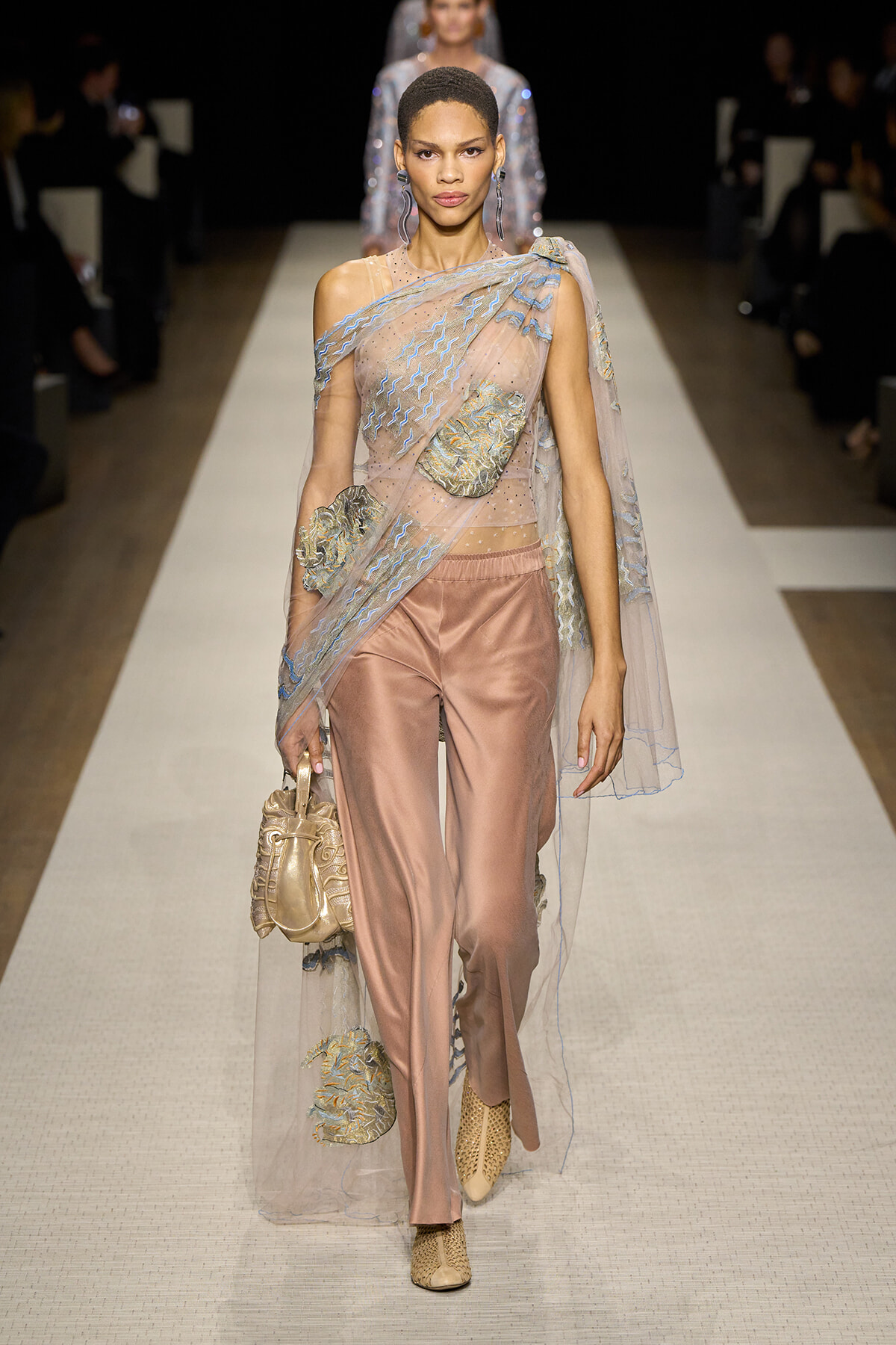 Model walks the runway in a sheer, embroidered cape over pale pink satin pants, paired with a metallic handbag and woven boots.