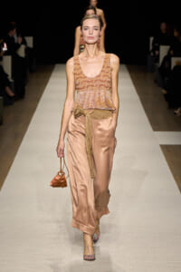 Model walking a fashion runway in a peach satin sleeveless top with fringe, matching wide-leg pants, and a fringe-tied belt, carrying a small orange purse with a chain