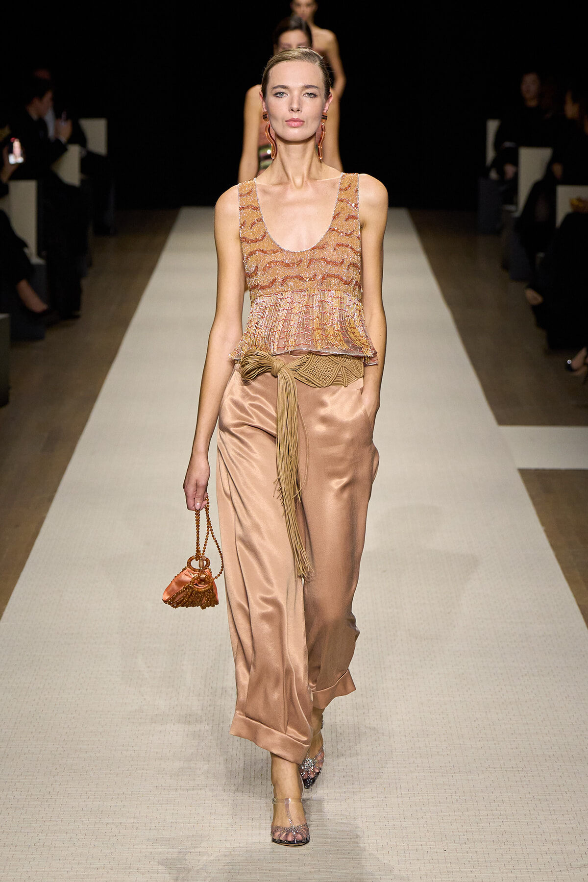 Model walking a fashion runway in a peach satin sleeveless top with fringe, matching wide-leg pants, and a fringe-tied belt, carrying a small orange purse with a chain
