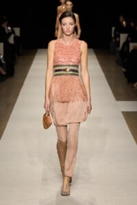 Model walks a runway in a peach sleeveless dress with fringe, cinched with a metallic belt, carrying a small beaded orange clutch.