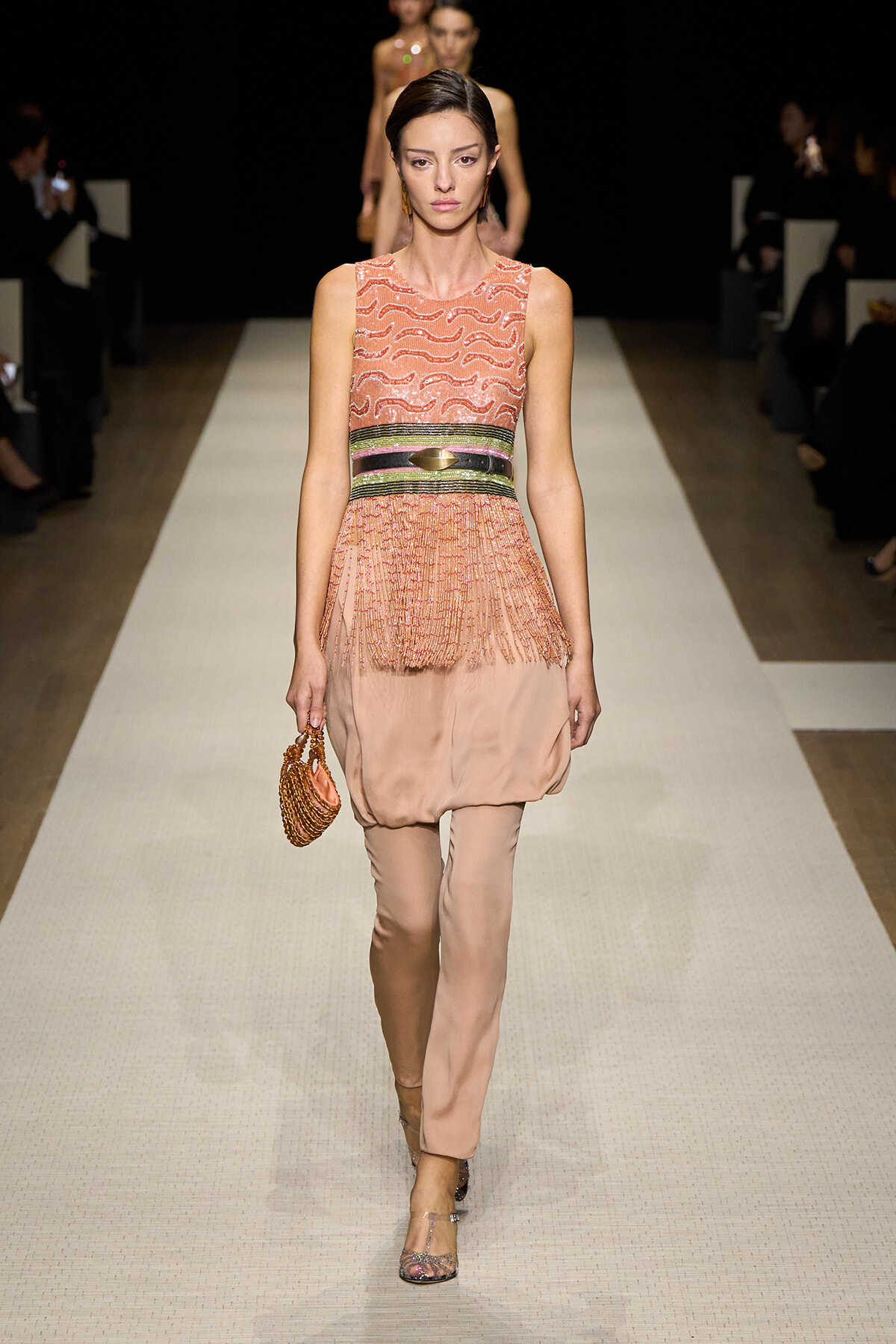 Model walks a runway in a peach sleeveless dress with fringe, cinched with a metallic belt, carrying a small beaded orange clutch.