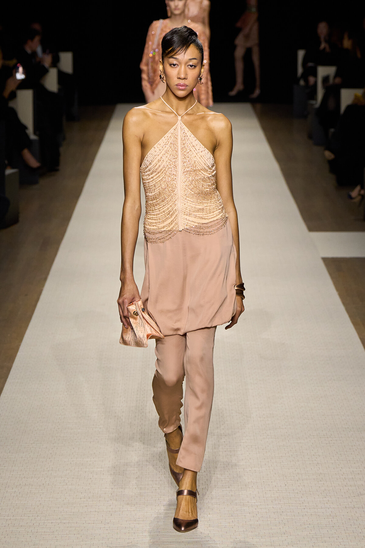 Model on runway wearing a peach beaded halter top with matching pants and a small clutch in hand.