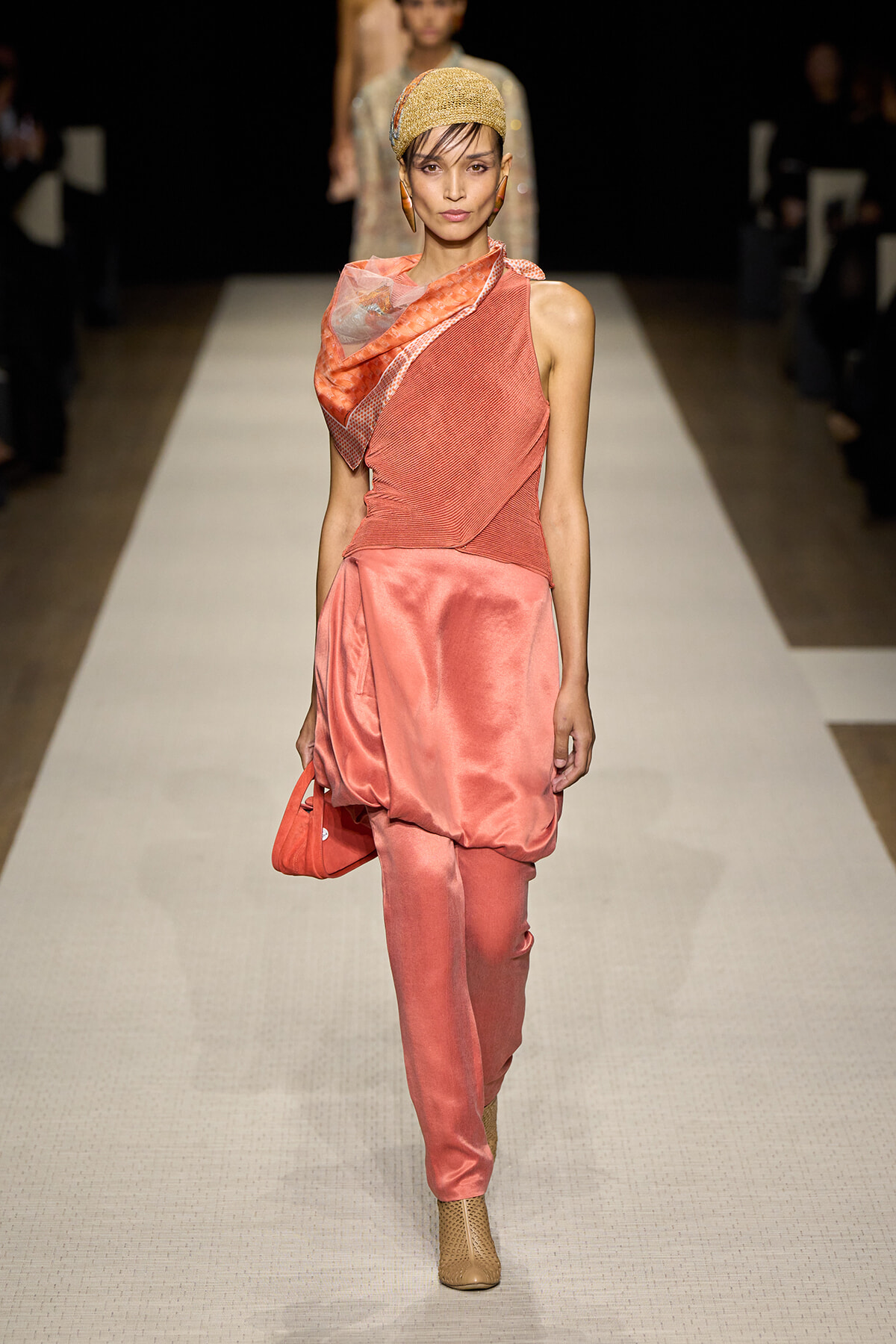 Runway model in coral satin ensemble: one-shoulder draped top with matching satin pants, tan boots, and a gold head wrap.