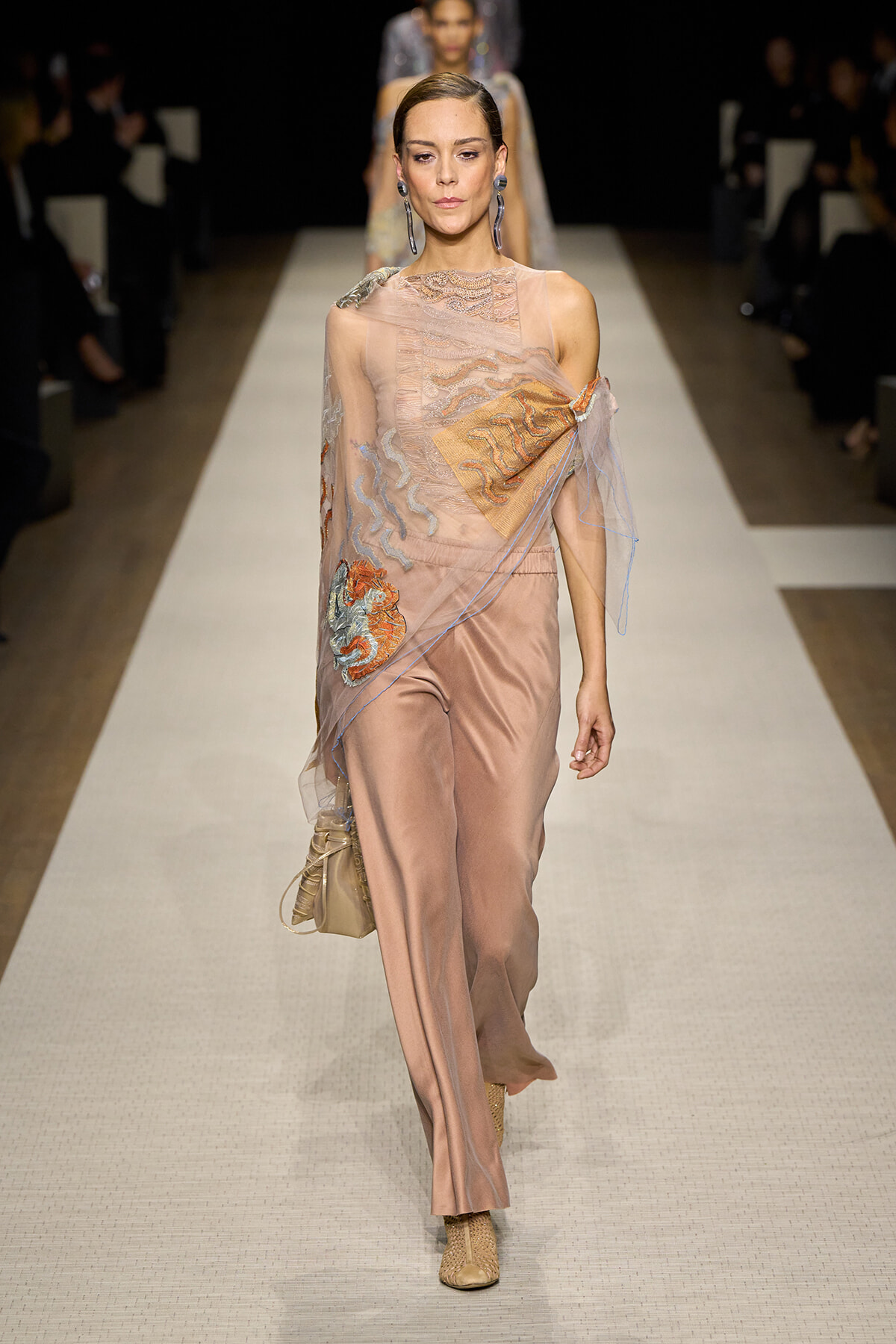 Model walking a fashion runway in a peach satin jumpsuit with sheer embroidered overlay and blue thread detailing, carrying a small beige handbag and wearing beige boots.
