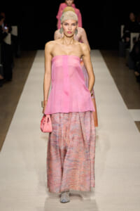 Model walking a pink strapless top over pink patterned maxi skirt on runway, accessorized with headwrap, earrings, and pink bag.