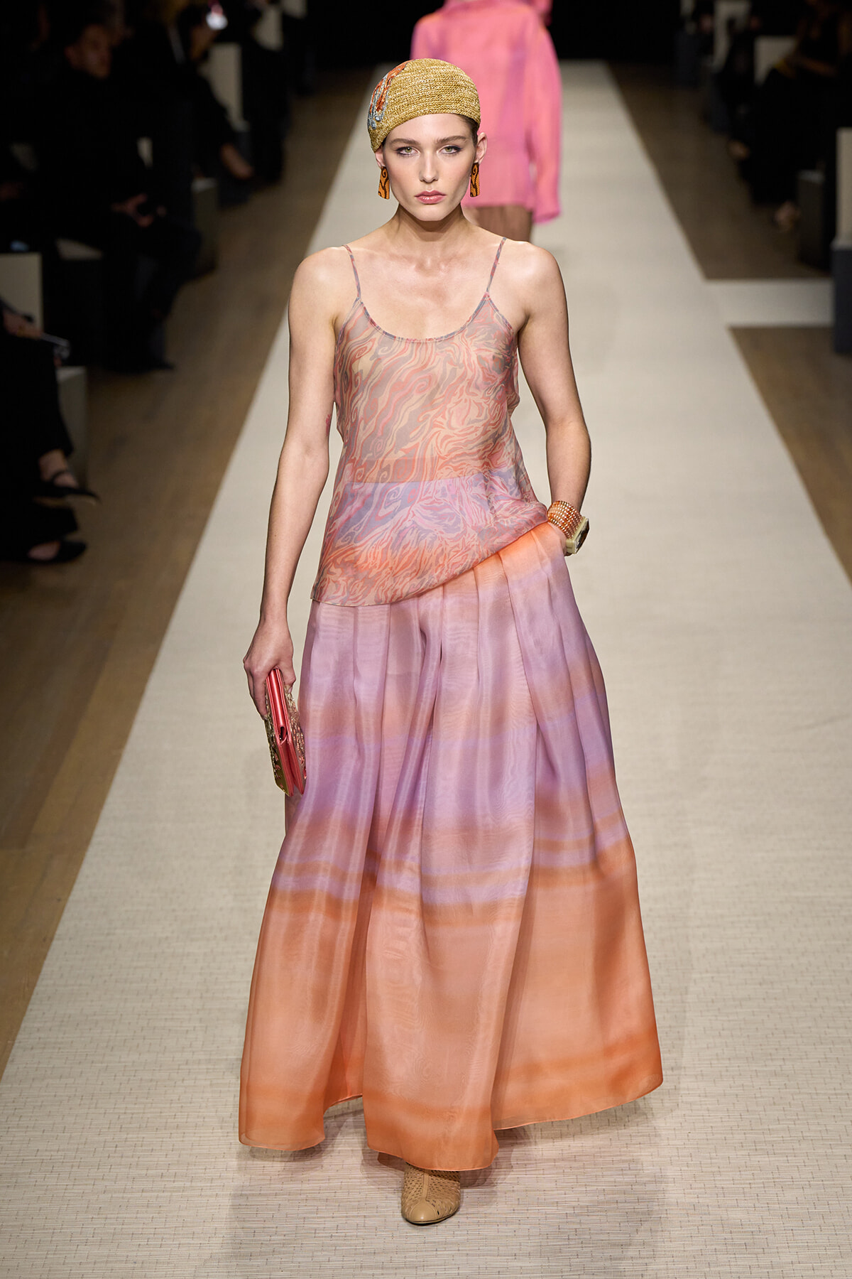 Model walking a runway in a peach–pink gradient satin skirt with a sheer patterned top, beige head wrap, earrings, bracelet, and a pink clutch.
