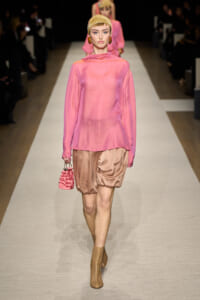 Runway model in a pink sheer blouse with a high neck, paired with bronze satin bubble shorts, tan perforated ankle boots, and a pink mini handbag, wearing a gold headwrap.